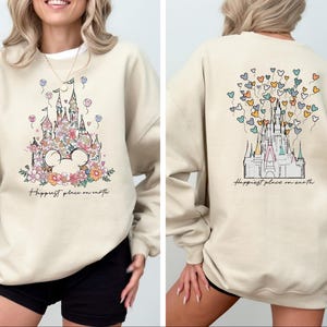 May include: A beige sweatshirt with a graphic of a floral castle and the text "Happiest place on earth". The castle is surrounded by colorful balloons in the shape of hearts.