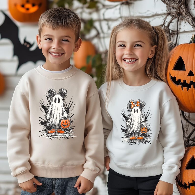 Peut inclure: Deux enfants portant des sweat-shirts sur le th&egrave;me d'Halloween. Le sweat-shirt beige pr&eacute;sente un fant&ocirc;me avec des oreilles de Mickey Mouse tenant une citrouille. Le sweat-shirt blanc a un fant&ocirc;me avec des oreilles de Minnie Mouse. Des citrouilles sont en arri&egrave;re-plan.