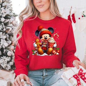 May include: Red Christmas sweatshirt featuring a cartoon image of Mickey Mouse wearing a Santa hat and scarf, holding a gingerbread man and a cup of hot chocolate.