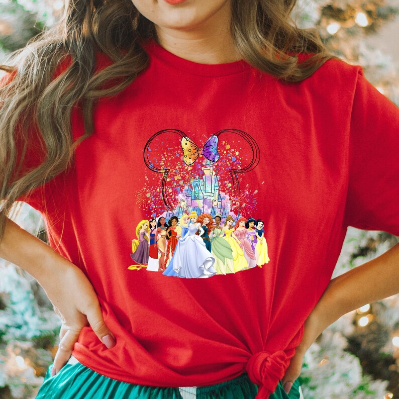 Puede incluir: Camiseta roja con un gr&aacute;fico colorido de princesas Disney frente al castillo de Cenicienta, orejas de Mickey Mouse y una mariposa. El gr&aacute;fico est&aacute; rodeado de confeti y fuegos artificiales.