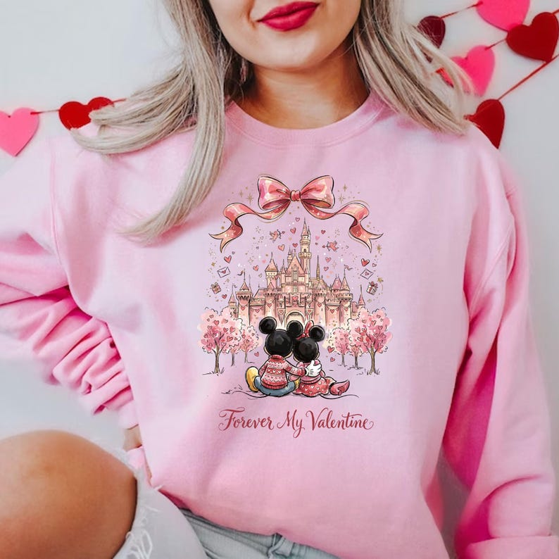 May include: Pink sweatshirt featuring a graphic of Mickey and Minnie Mouse in front of a castle, with a pink bow and the text "Forever My Valentine." The design includes pink trees and hearts, creating a romantic theme.