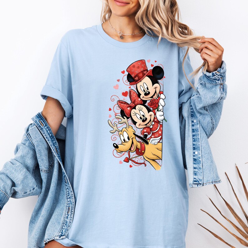 Puede incluir: Camiseta azul claro con Mickey Mouse, Minnie Mouse y Pluto con atuendos de San Valent&iacute;n. Mickey lleva un sombrero de copa, Minnie un lazo y Pluto un collar. El dise&ntilde;o incluye corazones rojos y una rosa.