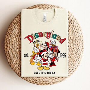 May include: A beige t-shirt with a vintage-style Disneyland logo featuring Mickey Mouse, Minnie Mouse, Donald Duck, Goofy, and Pluto in Christmas attire. The text reads "Disneyland est. 1955 California".