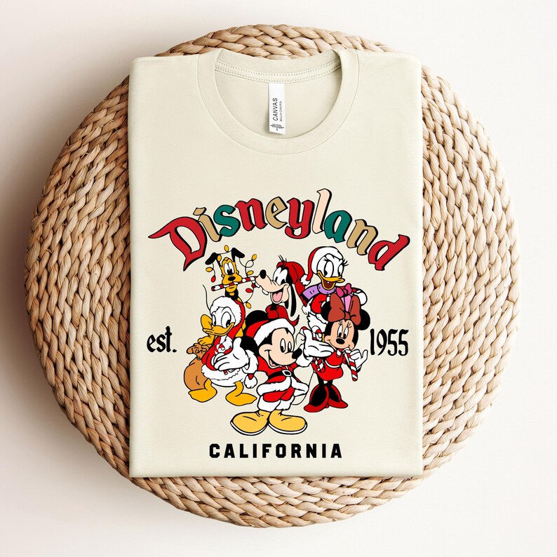 May include: A beige t-shirt with a vintage-style Disneyland logo featuring Mickey Mouse, Minnie Mouse, Donald Duck, Goofy, and Pluto in Christmas attire. The text reads "Disneyland est. 1955 California".