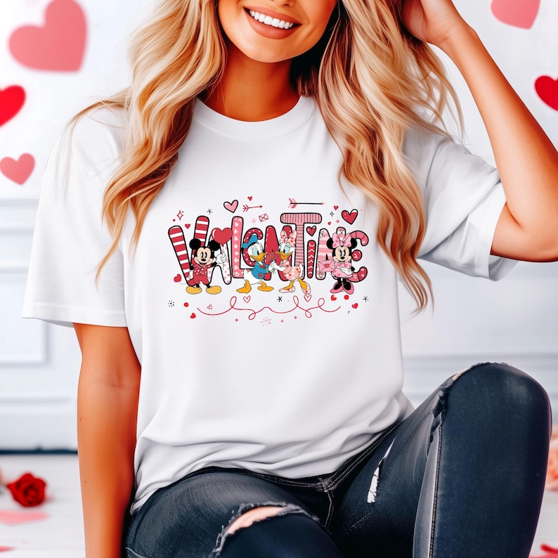 K&ouml;nnte beinhalten: Wei&szlig;es T-Shirt mit einem pinkfarbenen und roten Valentinstagsdesign mit Mickey Mouse, Minnie Mouse, Donald Duck und Goofy. Das Design sagt "Valentine" mit den Figuren, die in die Buchstaben integriert sind.