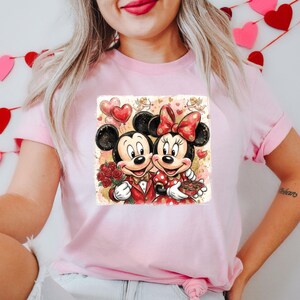May include: Pink t-shirt featuring a graphic of Mickey and Minnie Mouse in Valentine's Day attire. Mickey holds roses, Minnie holds chocolates, and both are smiling. The design includes heart-shaped balloons and a romantic background.