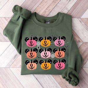May include: A green sweatshirt with a pattern of nine orange, pink, and peach pumpkins with Mickey Mouse ears and faces. The pumpkins are surrounded by black bats.