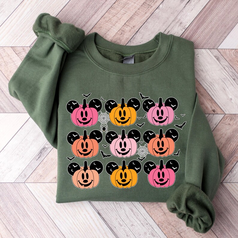 May include: A green sweatshirt with a pattern of nine orange, pink, and peach pumpkins with Mickey Mouse ears and faces. The pumpkins are surrounded by black bats.