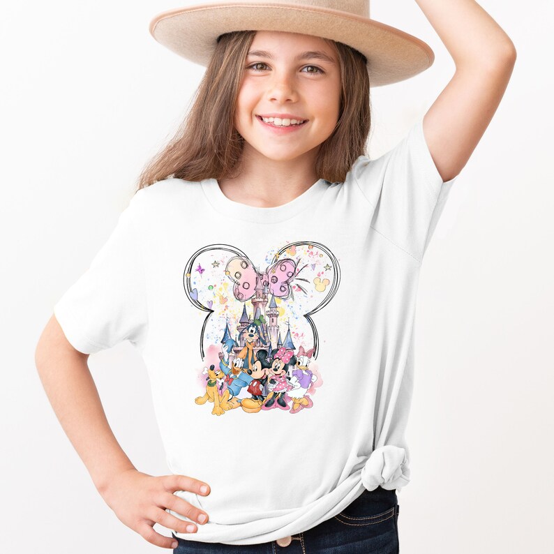 May include: White t-shirt with a colorful illustration of Mickey Mouse, Minnie Mouse, Goofy, Pluto, and Donald Duck in front of Cinderella's Castle. The illustration is surrounded by a Mickey Mouse ear shape and stars.