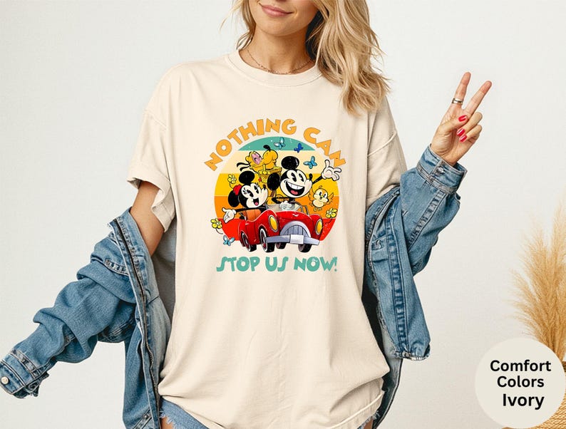 May include: Ivory-colored t-shirt featuring a colorful graphic of Mickey and Minnie Mouse in a red car, with the text "NOTHING CAN STOP US NOW!". The design includes a yellow sun, butterflies, and flowers.
