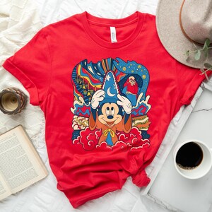 May include: Red t-shirt with a colorful graphic print of Mickey Mouse as a sorcerer. The graphic includes a blue hat with a crescent moon, a red and orange robe, and a background of swirling colors and clouds.