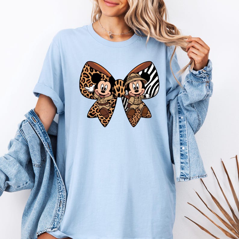 May include: Pale blue t-shirt with a large bow design featuring Mickey and Minnie Mouse in safari attire. The bow combines leopard print and zebra stripes. The cartoon characters are smiling and waving, creating a playful design.