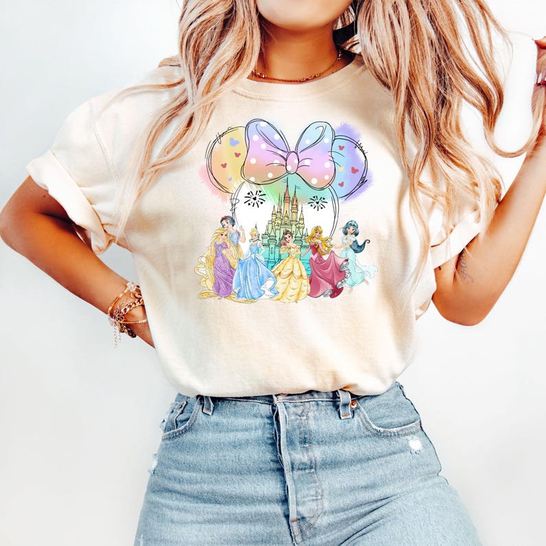 May include: A white t-shirt with a colorful graphic of Disney princesses and Cinderella's castle. The graphic is in the shape of Minnie Mouse ears with a pink bow.
