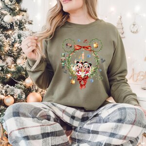 May include: Olive green sweatshirt featuring a festive Mickey and Minnie Mouse design. The graphic includes a wreath, bows, and a castle, with a nutcracker and ornaments. The long-sleeved top is perfect for the holiday season.