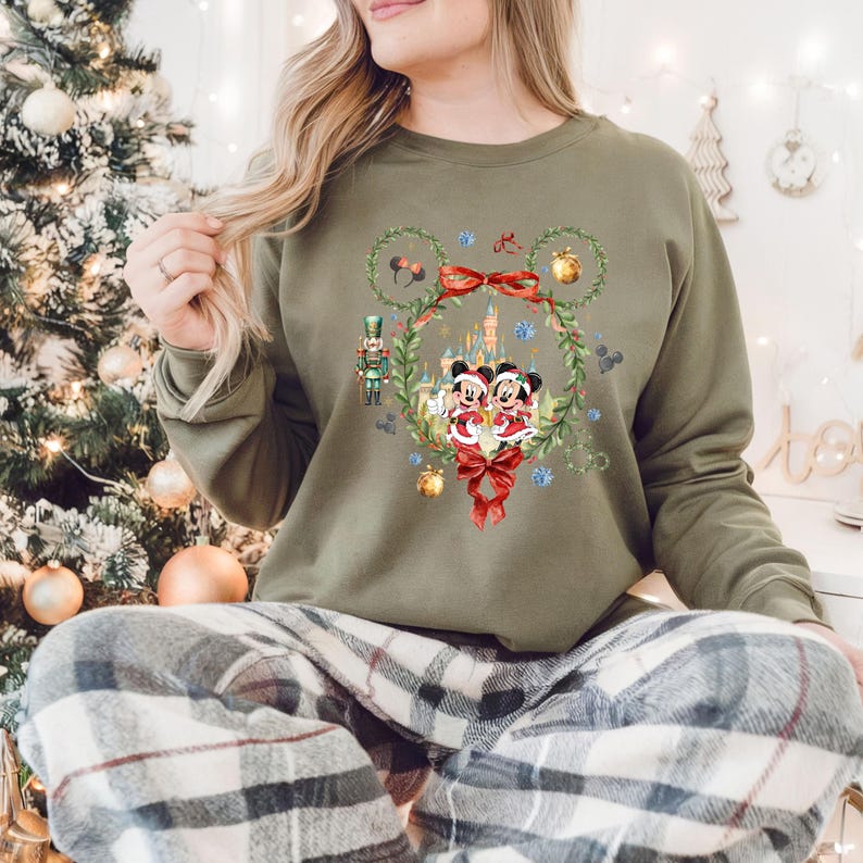 May include: Olive green sweatshirt featuring a festive Mickey and Minnie Mouse design. The graphic includes a wreath, bows, and a castle, with a nutcracker and ornaments. The long-sleeved top is perfect for the holiday season.