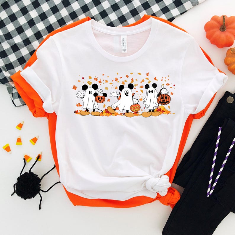 May include: White t-shirt with a graphic of four Mickey Mouse characters dressed as ghosts. The characters are holding pumpkins and walking through a field of orange leaves. The t-shirt is on a white background with a black and white checkered pattern in the background.