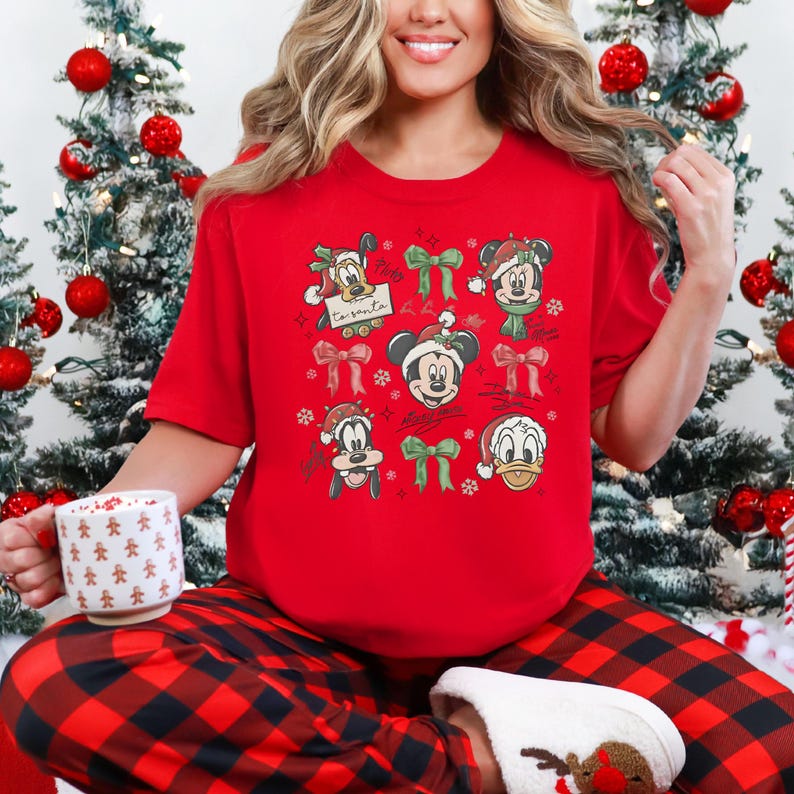 May include: A red Christmas-themed t-shirt featuring cartoon characters in Santa hats and green bows. The shirt has a festive design with Mickey Mouse, Donald Duck, and Goofy. The person is wearing red and black plaid pajama pants.