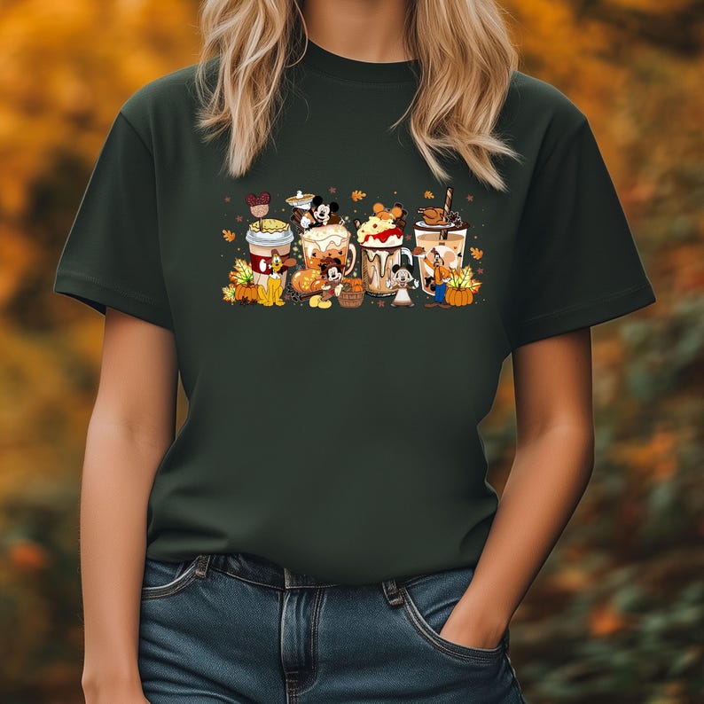 May include: Dark green t-shirt featuring a colorful graphic of Disney characters with fall-themed coffee drinks. The design includes Mickey Mouse, Minnie Mouse, and other characters, with pumpkins and autumn leaves. The shirt is a crew neck style.