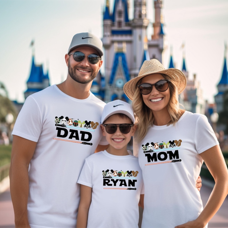 May include: Three people wearing white t-shirts with Disney character designs and text. The shirts say "DAD", "RYAN", and "MOM" with a lightsaber graphic. The background features a castle.