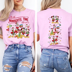 May include: A light pink t-shirt featuring a festive design with Disney characters. The front of the shirt displays Mickey, Minnie, Donald, Daisy, Goofy, and Pluto in holiday attire, with the text "The most WONDERFUL TIME of the year". The back has a vertical design of the characters.