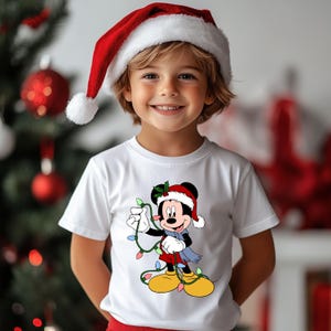 Puede incluir: Un ni&ntilde;o con una camiseta blanca con un personaje de dibujos animados de Mickey Mouse con gorro y bufanda de Pap&aacute; Noel, sosteniendo luces navide&ntilde;as. El fondo incluye un &aacute;rbol de Navidad y adornos.