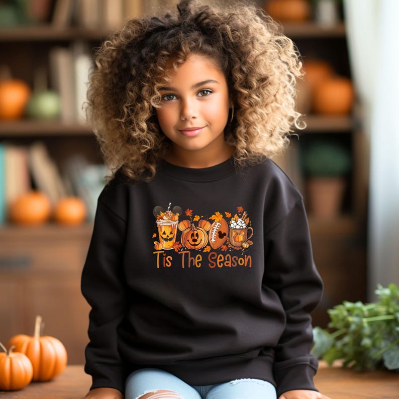 K&ouml;nnte beinhalten: Ein schwarzes Sweatshirt mit einem saisonalen Motiv, das ein Mickey Mouse-Getr&auml;nk, K&uuml;rbisse, einen Football und eine Tasse zeigt. Der Text "Tis The Season" ist darunter gedruckt. Das Sweatshirt wird von einer jungen Person getragen.