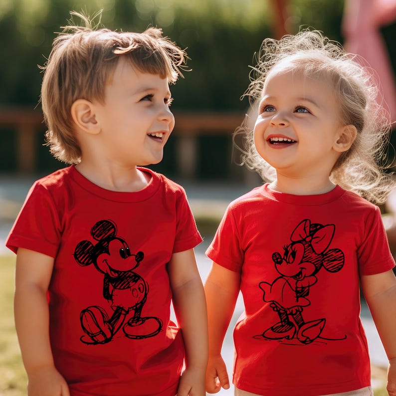 May include: Two children wearing red t-shirts with black and white cartoon images of Mickey Mouse and Minnie Mouse. The children are looking at each other and smiling.