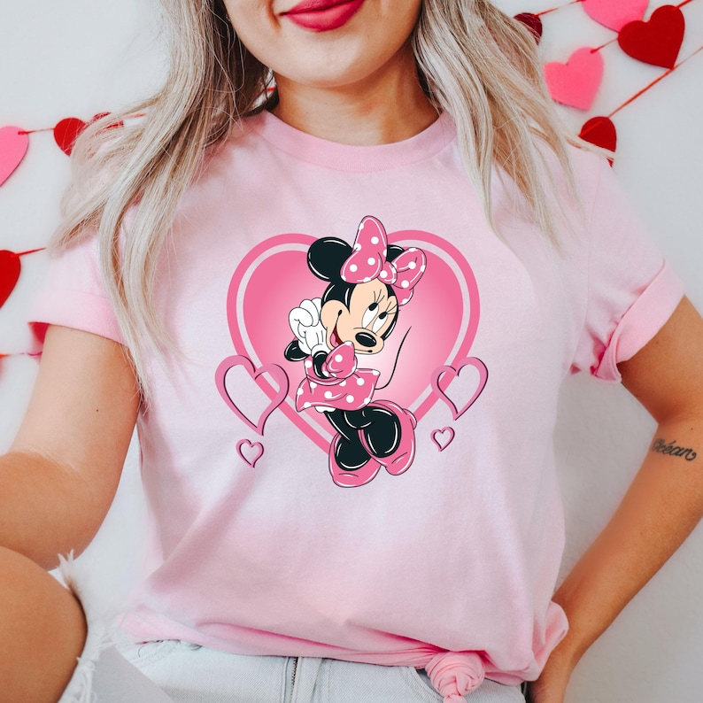 May include: Pink t-shirt with a graphic of Minnie Mouse in a pink heart with white polka dots. Minnie Mouse is wearing a pink dress with white polka dots and a pink bow.