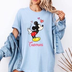 May include: Light blue t-shirt featuring a watercolor illustration of Mickey Mouse with hearts and the name "Thomas" in pink script. Mickey is in his classic pose, wearing red shorts and yellow shoes.