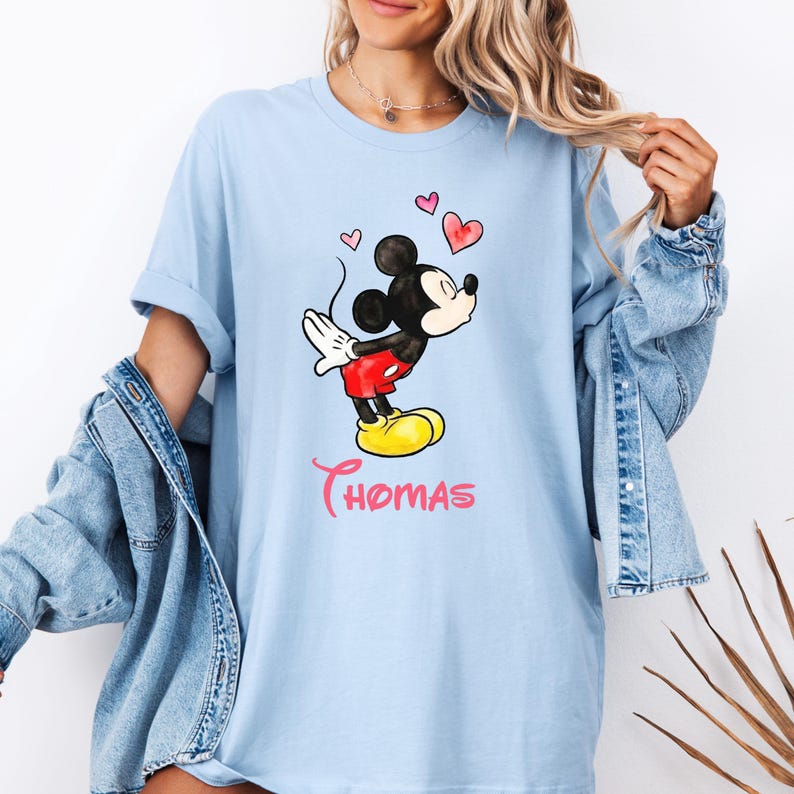 May include: Light blue t-shirt featuring a watercolor illustration of Mickey Mouse with hearts and the name "Thomas" in pink script. Mickey is in his classic pose, wearing red shorts and yellow shoes.