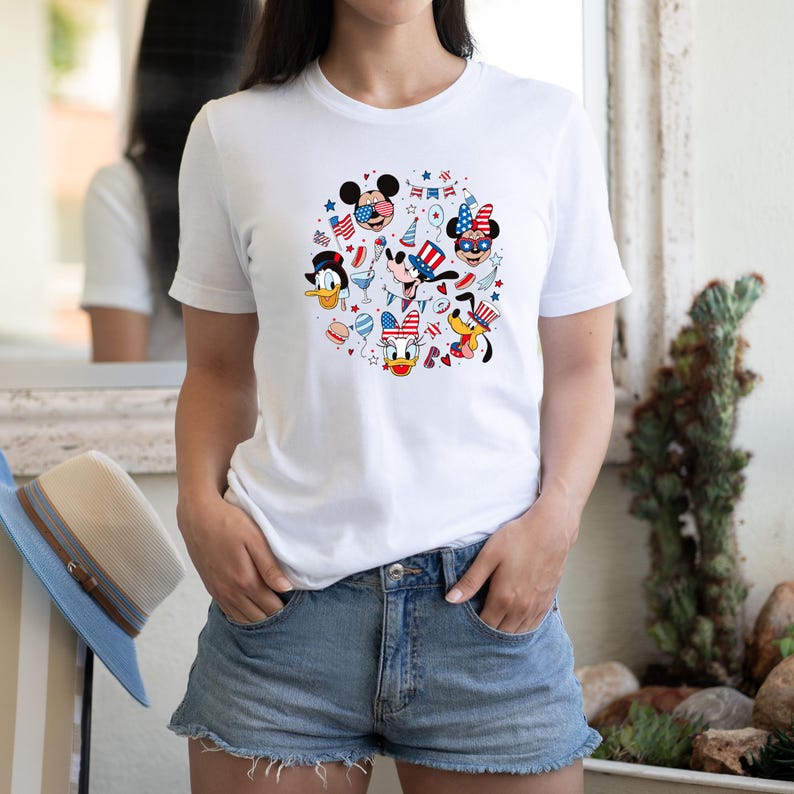 May include: White t-shirt featuring a circular design with cartoon characters Mickey Mouse, Minnie Mouse, Donald Duck, Goofy, and Pluto, all in patriotic attire. The design includes red, white, and blue stars, stripes, and festive elements.