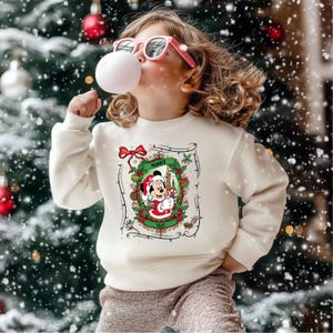 Może przedstawiać: Kremowa bluza świąteczna z Myszka Minnie. Grafika przedstawia Minnie w stroju Świętego Mikołaja, otoczoną świątecznym wieńcem i napisem "Minnie". Tło obejmuje choinkę z ozdobami i padającym śniegiem.