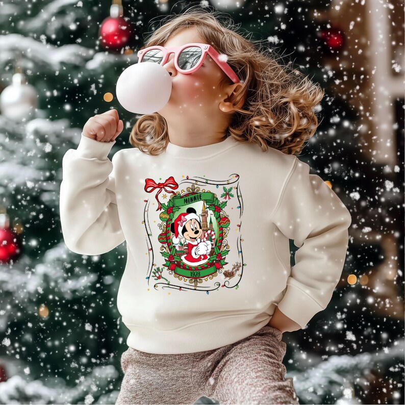 Może przedstawiać: Kremowa bluza świąteczna z Myszka Minnie. Grafika przedstawia Minnie w stroju Świętego Mikołaja, otoczoną świątecznym wieńcem i napisem "Minnie". Tło obejmuje choinkę z ozdobami i padającym śniegiem.