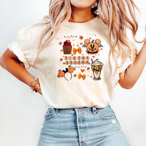 Puede incluir: Camiseta color crema con un dise&ntilde;o tem&aacute;tico de Halloween. El dise&ntilde;o incluye una calabaza con orejas de rat&oacute;n, una magdalena, lazos y una bebida de caf&eacute;. Tambi&eacute;n aparece el texto "Tis the Season".