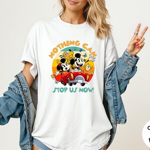 May include: White t-shirt featuring a colorful graphic of Mickey and Minnie Mouse in a red car, with the text "NOTHING CAN STOP US NOW!". The design includes a yellow, orange, and blue sunset background. The shirt is a comfort colors white.