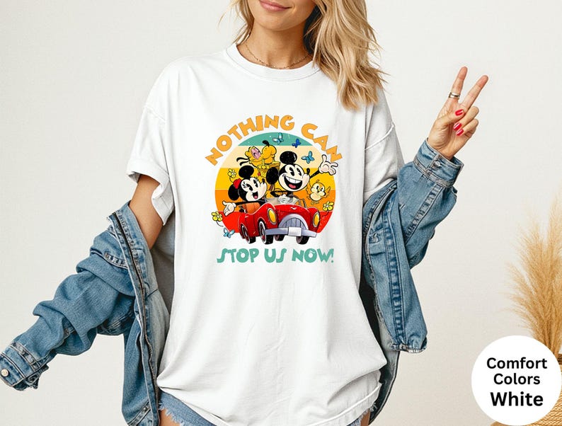 May include: White t-shirt featuring a colorful graphic of Mickey and Minnie Mouse in a red car, with the text "NOTHING CAN STOP US NOW!". The design includes a yellow, orange, and blue sunset background. The shirt is a comfort colors white.