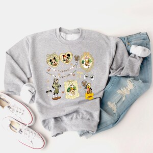 May include: A light gray sweatshirt featuring cartoon character graphics in gold frames and heart shapes. The text "ANIMAL KINGDOM" is printed on the sweatshirt. The sweatshirt is paired with blue jeans and white sneakers.