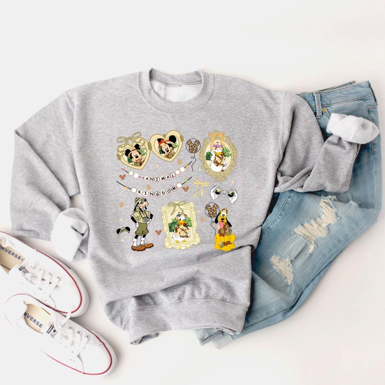 May include: A light gray sweatshirt featuring cartoon character graphics in gold frames and heart shapes. The text "ANIMAL KINGDOM" is printed on the sweatshirt. The sweatshirt is paired with blue jeans and white sneakers.