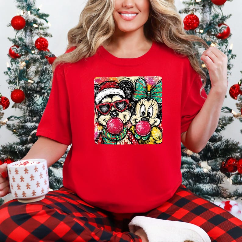 May include: Red t-shirt featuring a colorful graphic of Mickey and Minnie Mouse blowing bubble gum. Mickey wears a Santa hat and sunglasses. Minnie has a green bow. The shirt is worn with red and black plaid pants.