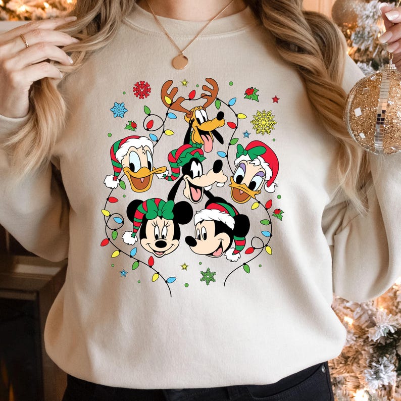 May include: A beige sweatshirt with a Christmas-themed design featuring Mickey Mouse, Minnie Mouse, Donald Duck, Goofy, and Pluto. The characters are surrounded by colorful Christmas lights and snowflakes.