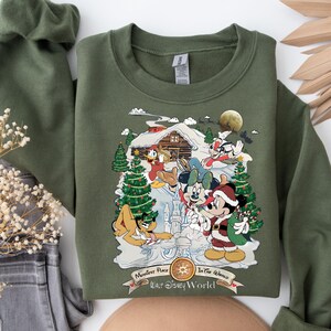 Pu&ograve; includere: Felpa verde con un design a tema natalizio con Topolino, Minnie Mouse, Paperino, Pippo e Pluto. Il design include una baita, un albero di Natale e il testo "Happiest Place on Earth, Walt Disney World".