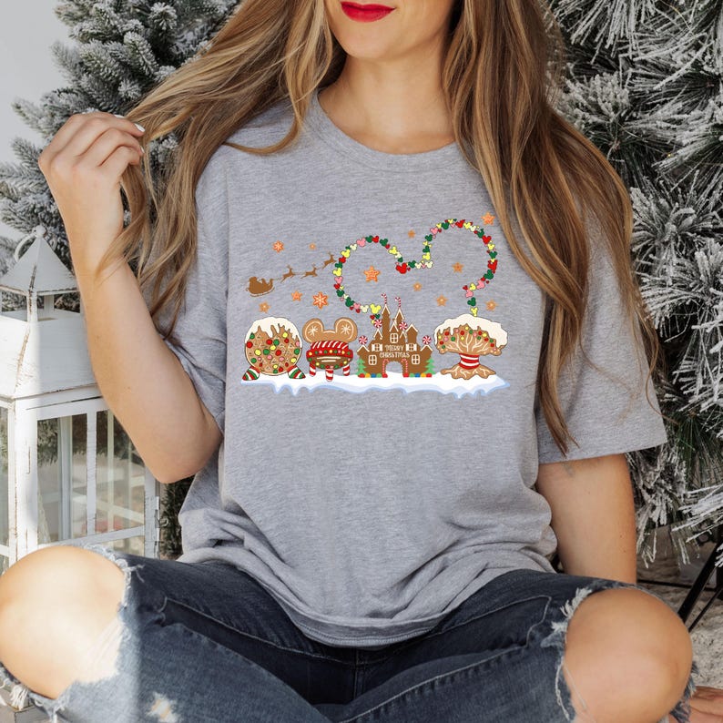 May include: A grey t-shirt with a gingerbread themed Christmas design featuring a Mickey Mouse head, a gingerbread castle, and a gingerbread tree. The design includes the text "Merry Christmas".