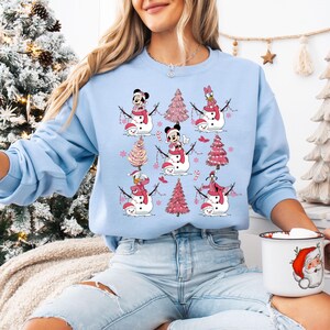 May include: Pale blue sweatshirt with a repeating pattern of snowmen featuring Disney characters, pink Christmas trees, and candy canes. The snowmen wear Santa hats and scarves. A festive holiday design.