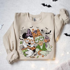 May include: A beige sweatshirt with a Halloween-themed graphic featuring Mickey Mouse and Donald Duck wearing heart-shaped sunglasses. The graphic includes spiderwebs, bats, and candy.
