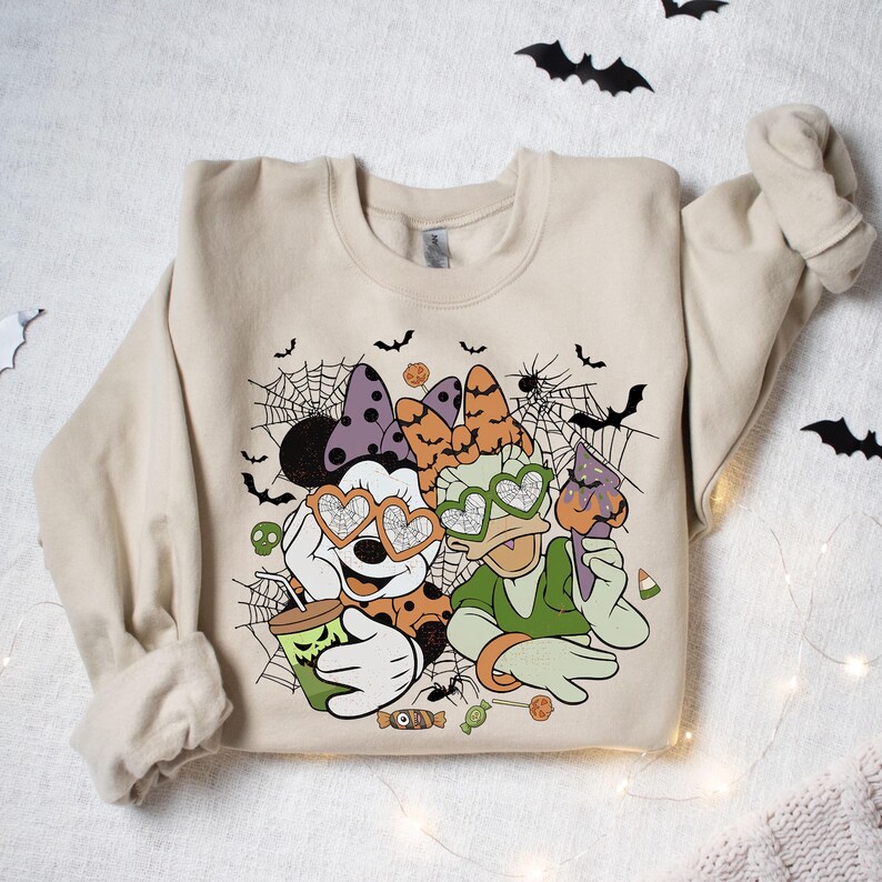 May include: A beige sweatshirt with a Halloween-themed graphic featuring Mickey Mouse and Donald Duck wearing heart-shaped sunglasses. The graphic includes spiderwebs, bats, and candy.