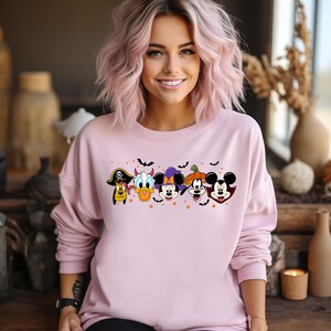 Op de afbeelding: Roze sweatshirt met een grafische print van Mickey Mouse, Minnie Mouse, Donald Duck, Goofy en Pluto in Halloween-kostuums. De personages zijn in een rij gerangschikt en hebben vleermuizen die boven hen vliegen.
