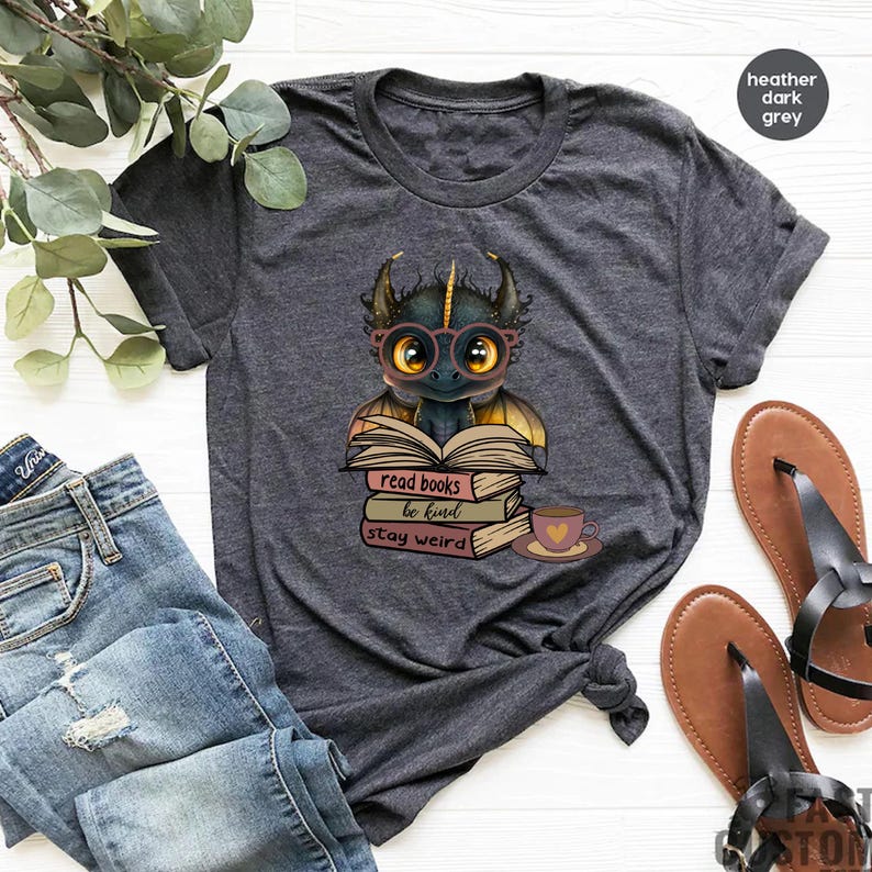 May include: A dark heather grey t-shirt with a cartoon dragon wearing glasses and sitting on a stack of books. The books have the text "read books be kind stay weird".