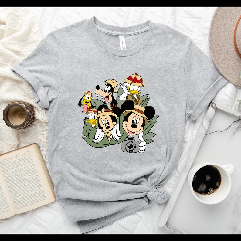 May include: A grey t-shirt featuring a graphic print of Mickey Mouse, Minnie Mouse, Goofy, Donald Duck and Pluto dressed in safari gear. The characters are surrounded by green leaves.