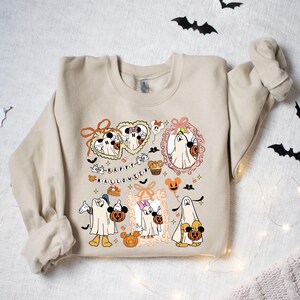 May include: Beige sweatshirt featuring a Halloween-themed graphic. The design includes cartoon characters dressed as ghosts, pumpkins, bats, and the text "HAPPY HALLOWEEN". The graphic is in shades of orange, black, and white.