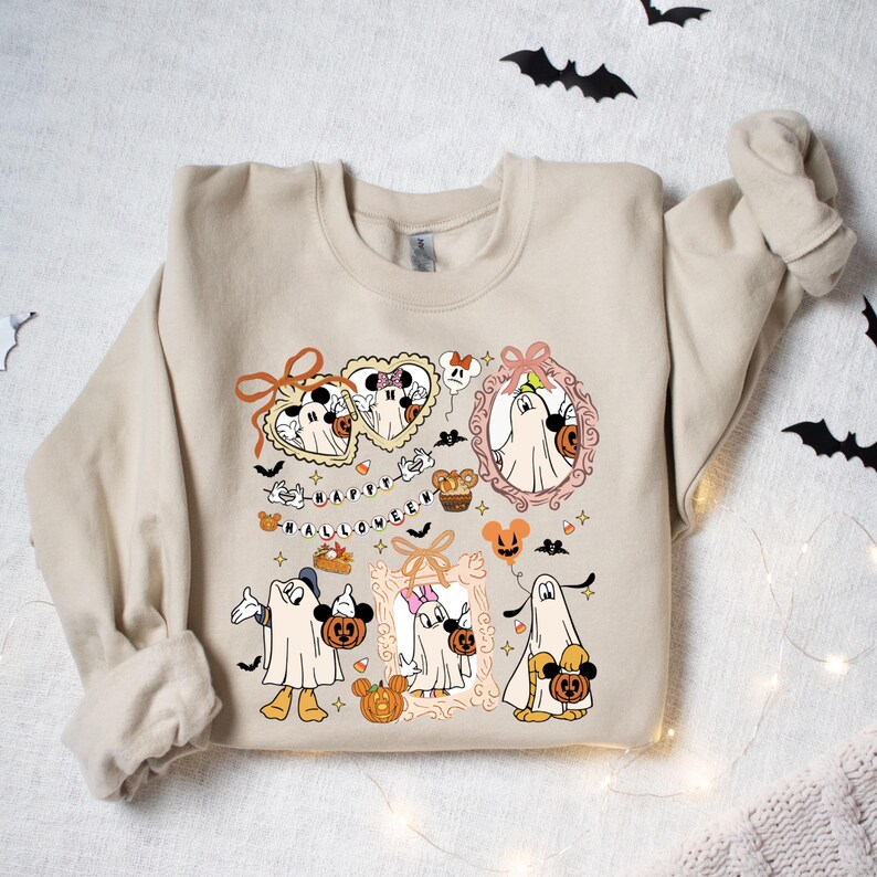 May include: Beige sweatshirt featuring a Halloween-themed graphic. The design includes cartoon characters dressed as ghosts, pumpkins, bats, and the text "HAPPY HALLOWEEN". The graphic is in shades of orange, black, and white.